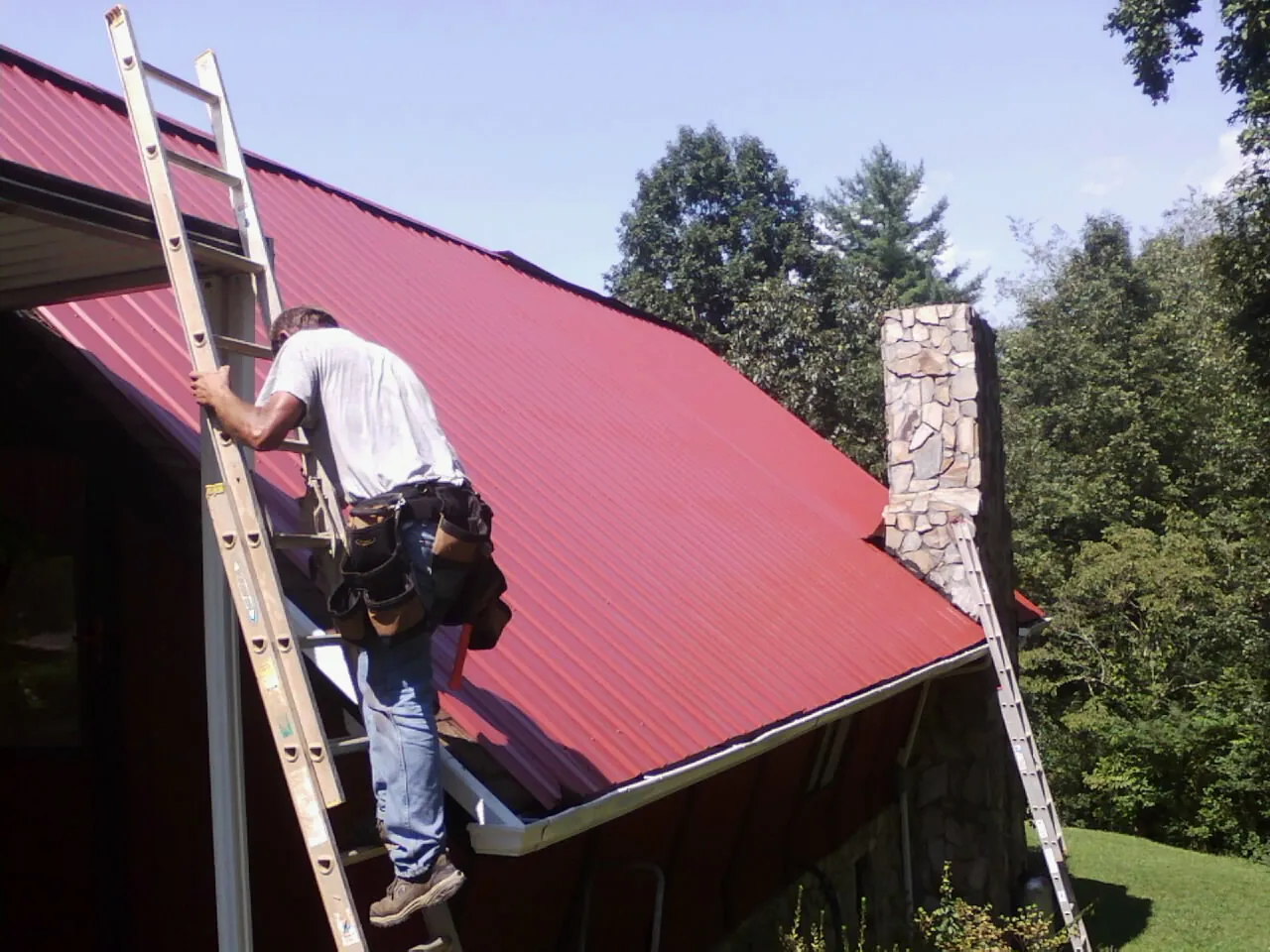 Roofer on a ladder beside a red metal roof and stone chimney — Soffit & Fascia Repair by Roofing Brothers in Appleton